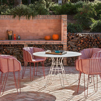 "Soft pink chairs with sleek white table against a textured brick wall and natural wood accents in a warm outdoor setting."