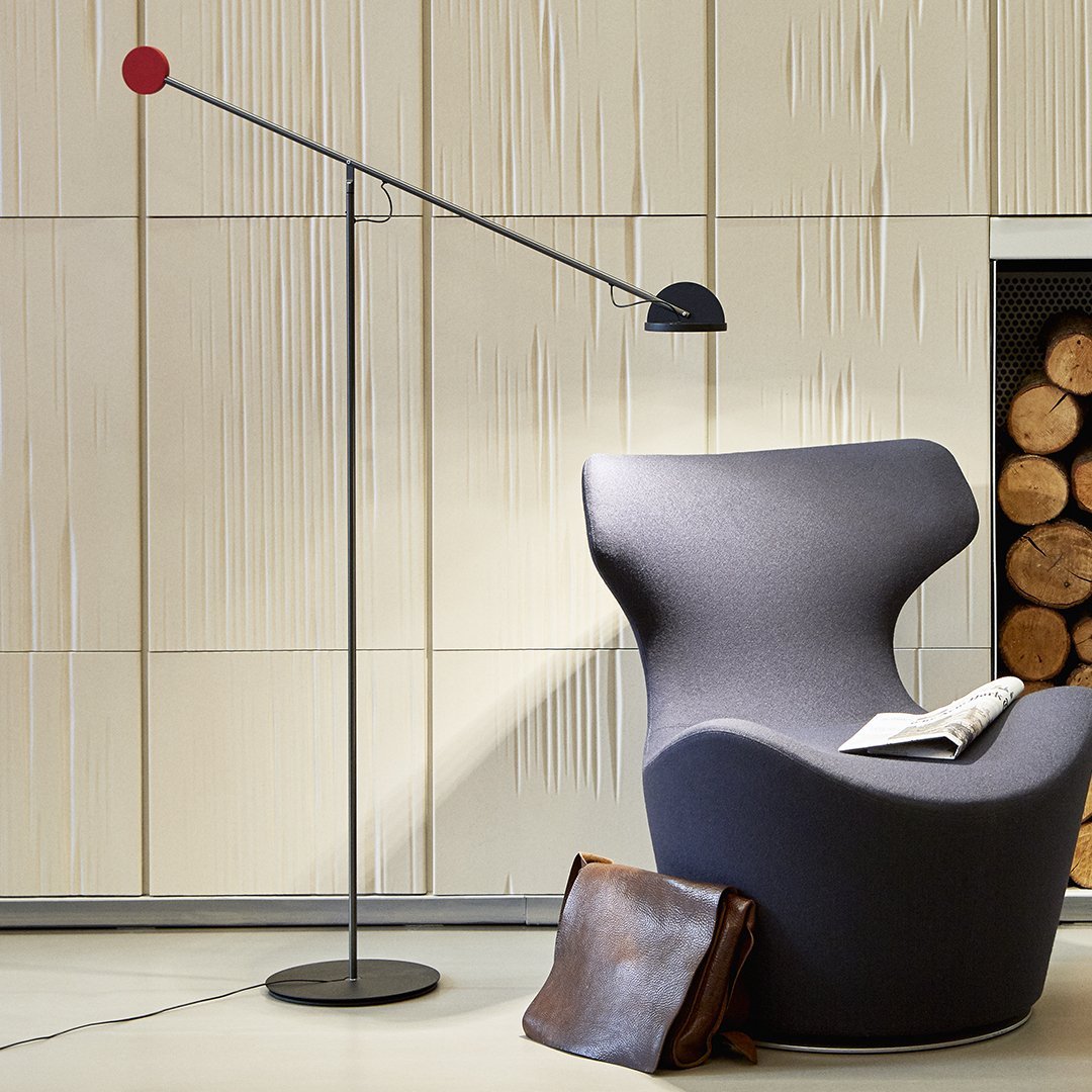 Modern gray chair with a sleek black floor lamp featuring a red accent, set against a textured light wood backdrop.