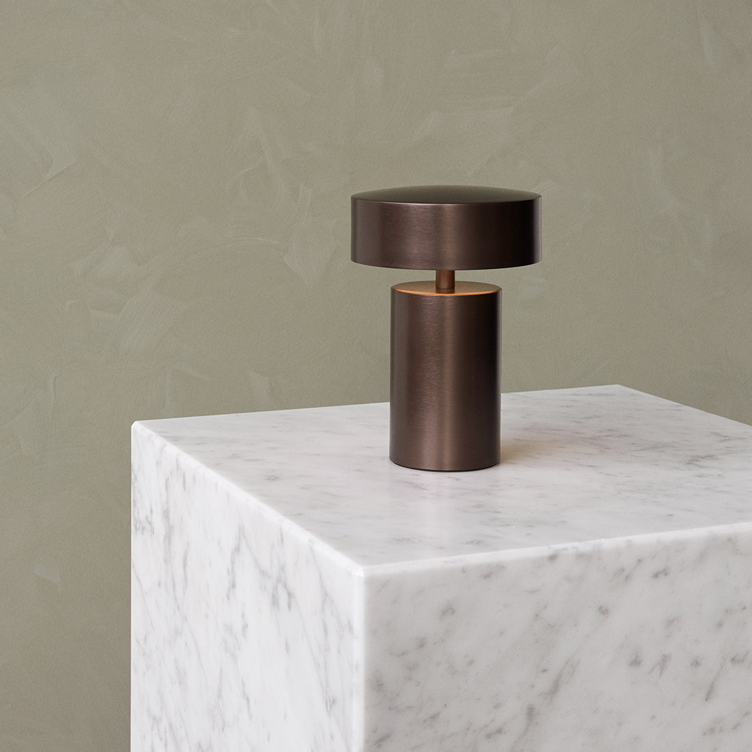 Bronze lamp with minimalist design on a white marble pedestal, set against a muted green textured wall.