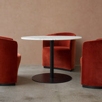 Round marble table with black base, surrounded by two plush burnt orange chairs against a neutral backdrop.
