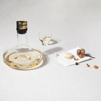 Clear glass decanter and cup with golden accents; beige macaron and brown decorative balls on white napkin.