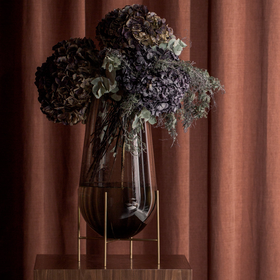 "Elegant black glass vase with dark purple and green dried flowers against warm rust-colored curtains."