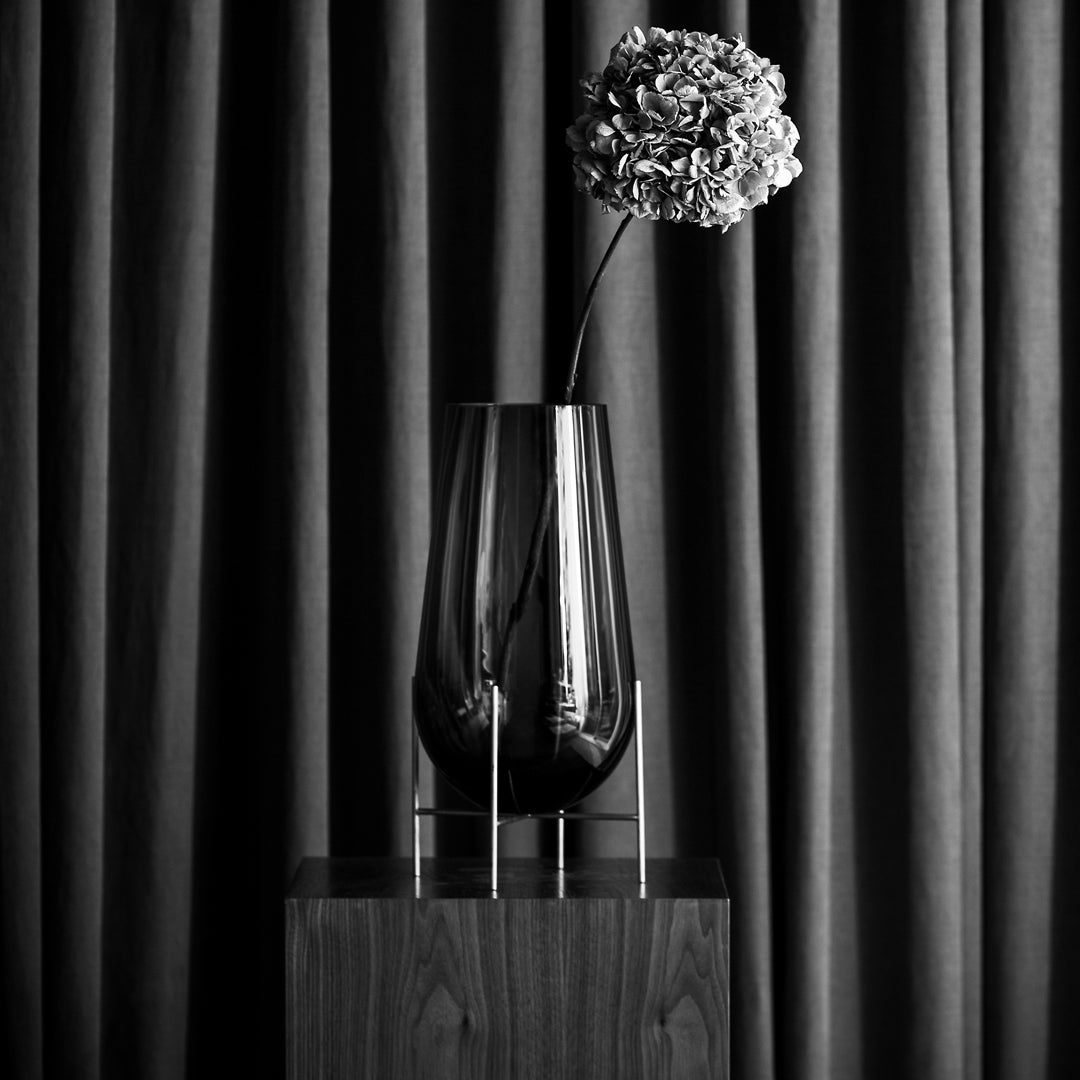 Black glass vase with a modern design, featuring a delicate flower against textured dark gray curtains.