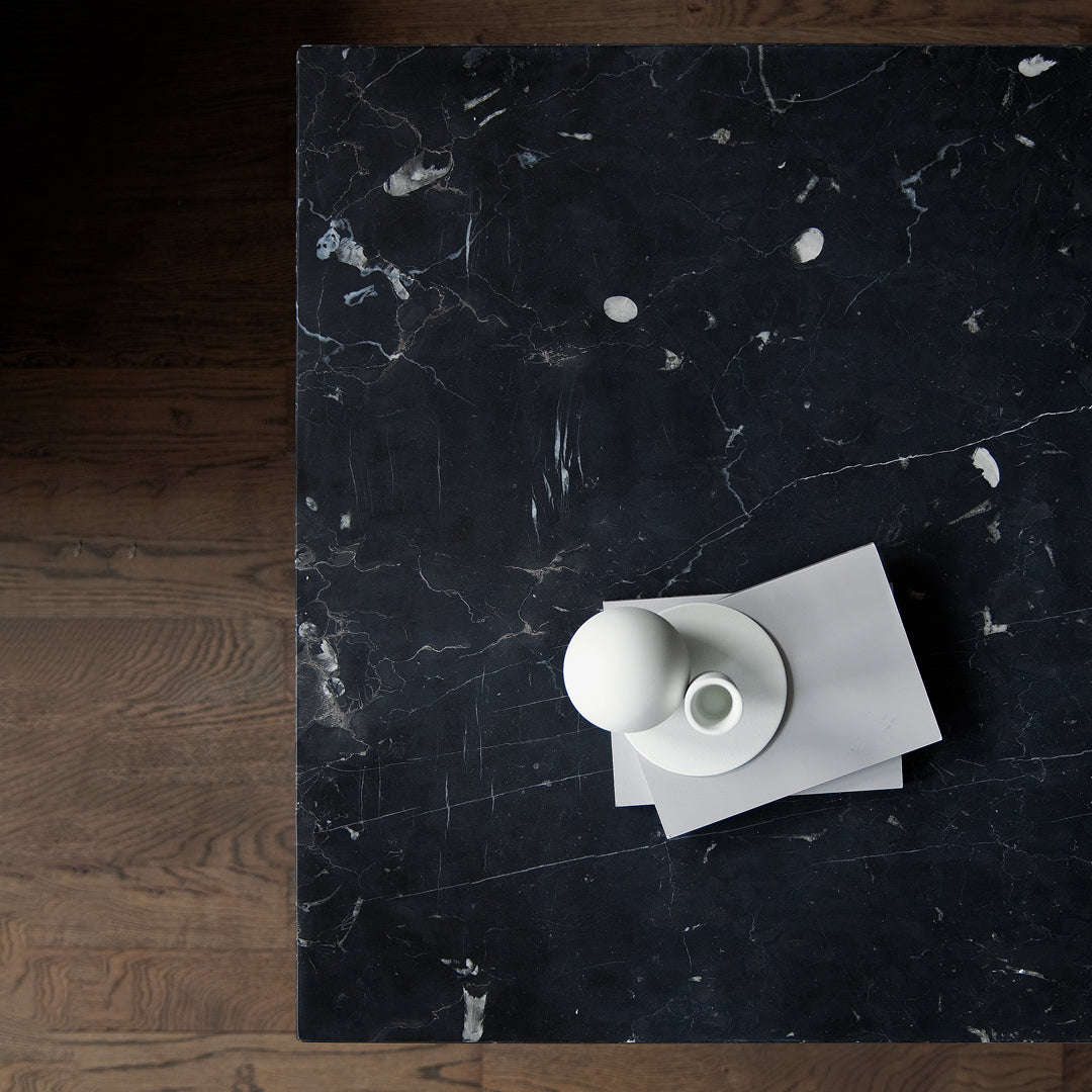 "Dark marble table with white veining, featuring a minimalist white vase and grey stack of papers."