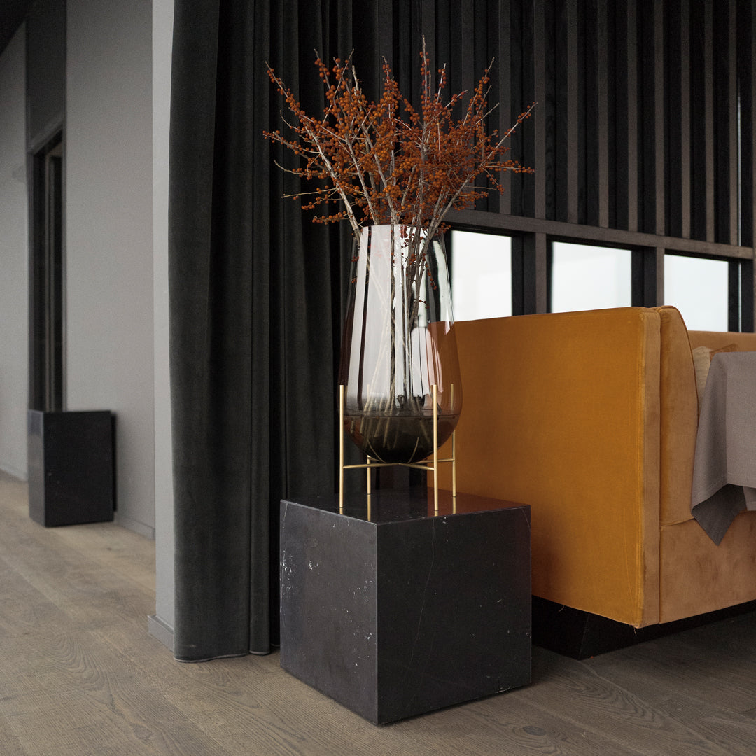 Clear glass vase with dried orange foliage, set on a sleek black pedestal next to a warm brown sofa. Modern decor accents.