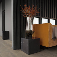 Clear glass vase with dried orange foliage, set on a sleek black pedestal next to a warm brown sofa. Modern decor accents.