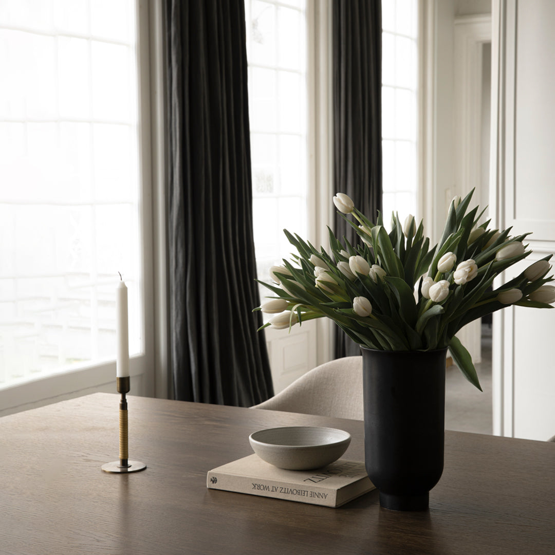 "Elegant black vase with white tulips, gray bowl, and gold candle holder on a wooden table with dark curtains."