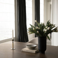 "Elegant black vase with white tulips, gray bowl, and gold candle holder on a wooden table with dark curtains."