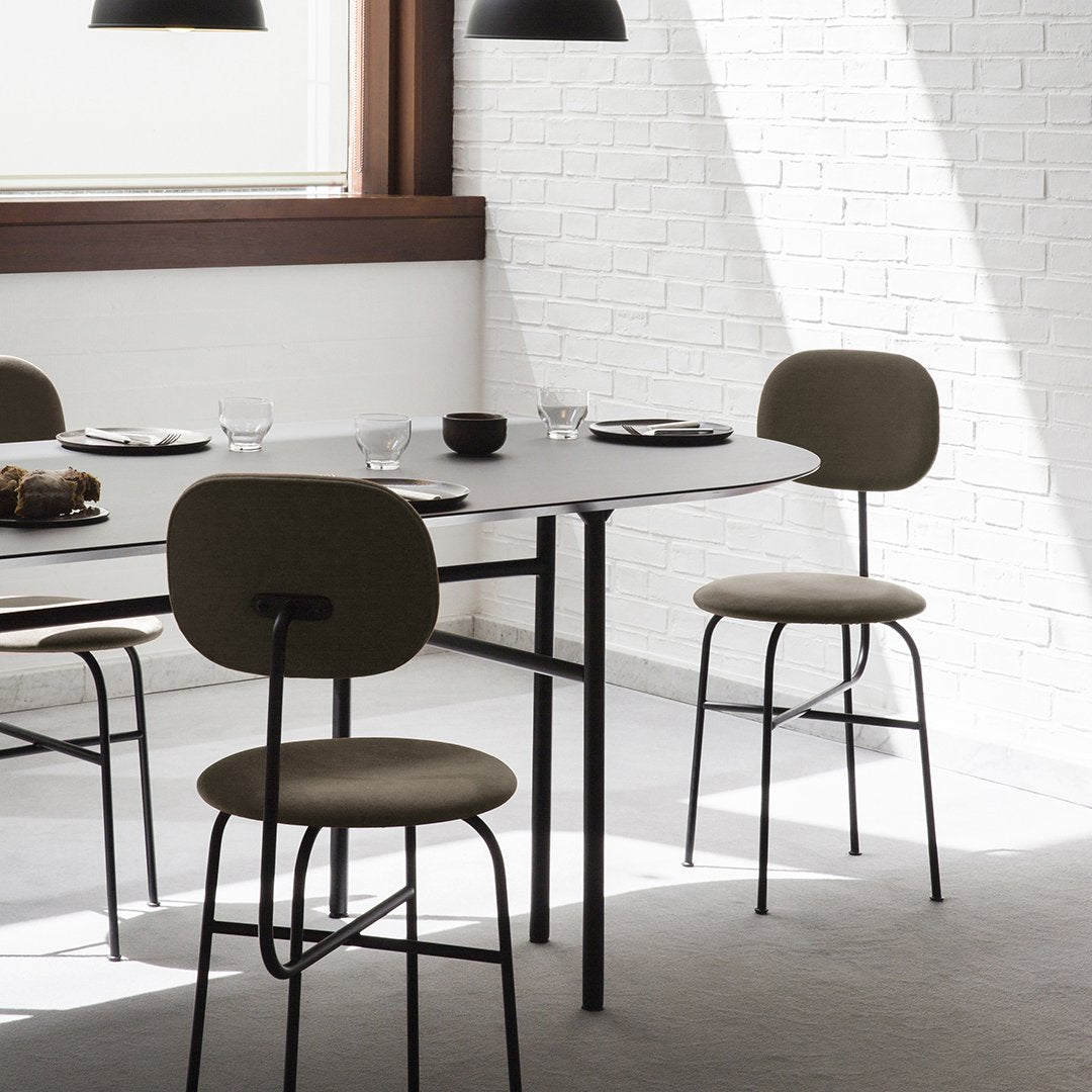Modern dining room with beige upholstered chairs and a gray round table against a white brick wall. Minimalist design elements.
