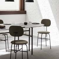Modern dining room with beige upholstered chairs and a gray round table against a white brick wall. Minimalist design elements.
