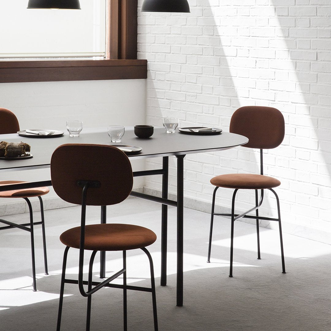 Stylish round table with black legs, paired with rich brown cushioned chairs against a white brick wall.