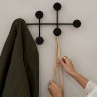 Black wall hook with circular accents, showcasing an olive green coat and beige ribbons against a neutral wall.