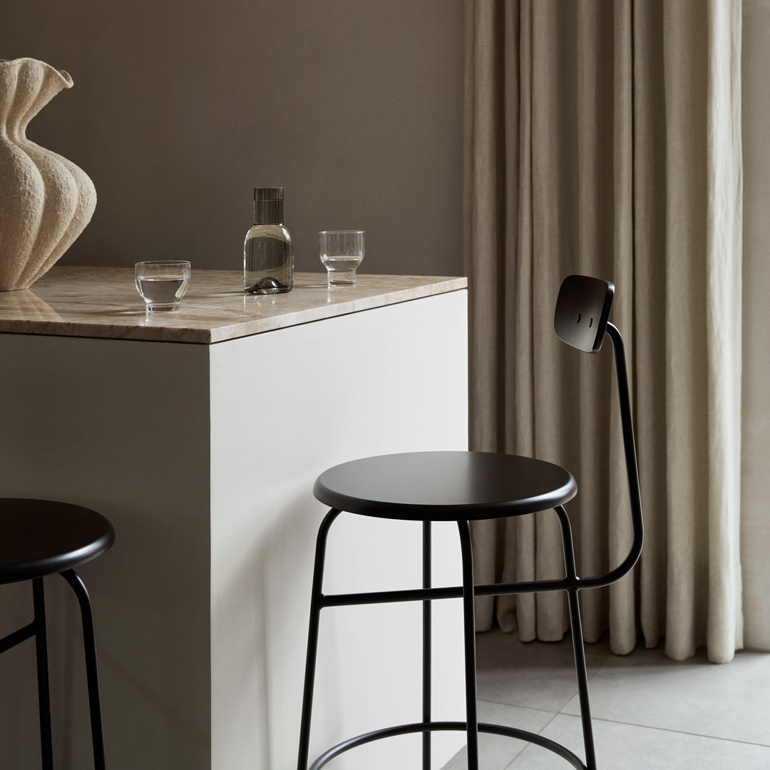 Black minimalist bar stools on a light wooden countertop with soft beige curtains in a neutral-toned setting.