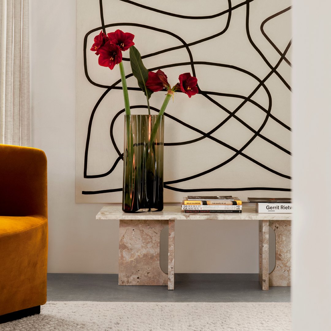 "Bright red flowers in a black vase against a creamy backdrop with abstract black lines, complemented by a golden orange couch."
