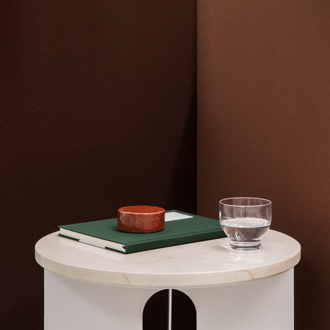 White round side table with a marble top, green book, brown wooden bowl, and clear glass on a rich brown background.