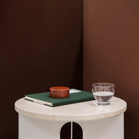 White round side table with a marble top, green book, brown wooden bowl, and clear glass on a rich brown background.