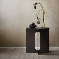 Black modern side table with a curved cutout, showcasing a sleek design against a neutral wall and warm wooden floor.
