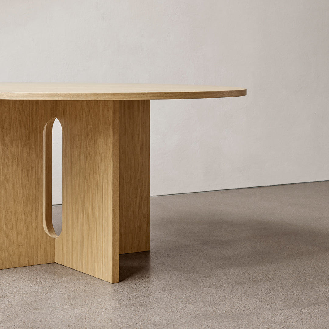 Natural wood finish table with a smooth oval top and minimalist cut-out pattern, set against a gray-toned floor.