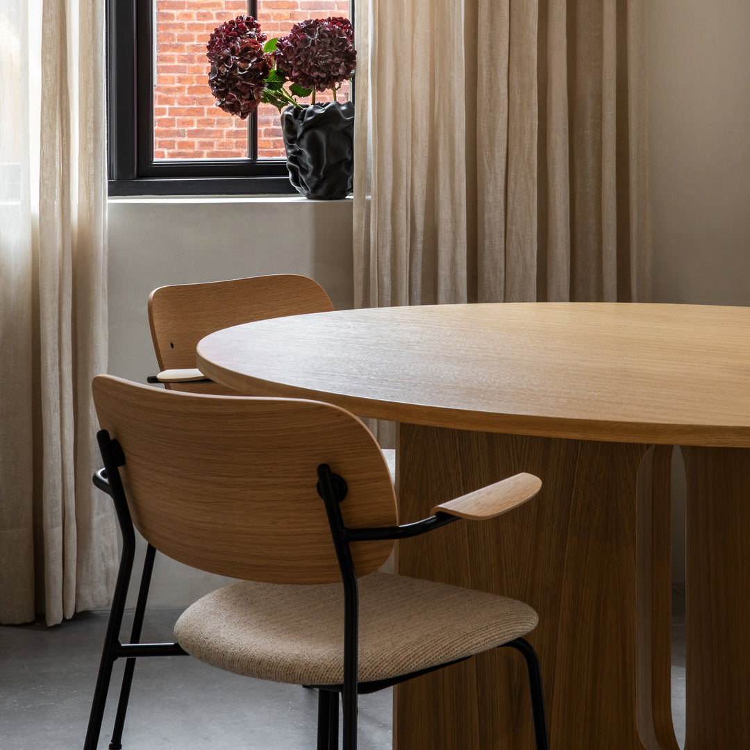 Natural wood table with light beige chairs and soft cream curtains, accentuated by deep purple flowers in a vase.