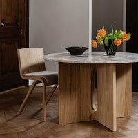 Round marble-top table with warm beige wood base, surrounded by a light wood chair and vibrant orange flowers.