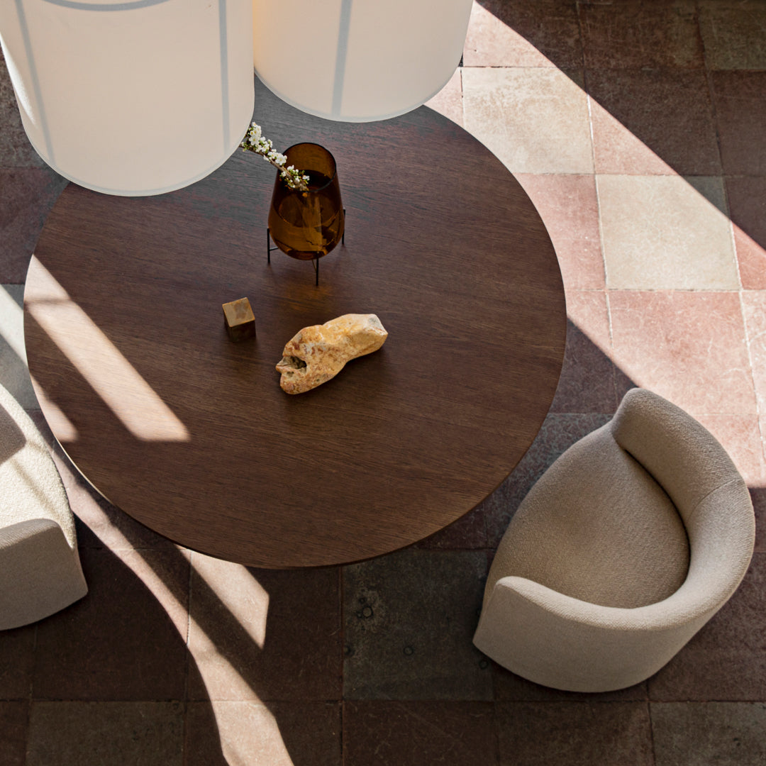 Round wooden table on patterned tiles, surrounded by beige armchairs and natural decor highlights.