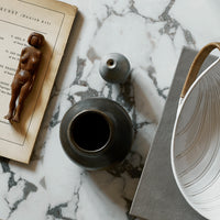 Dark ceramic vase and white patterned bowl on marble surface with book and wooden figure, showcasing neutral tones.