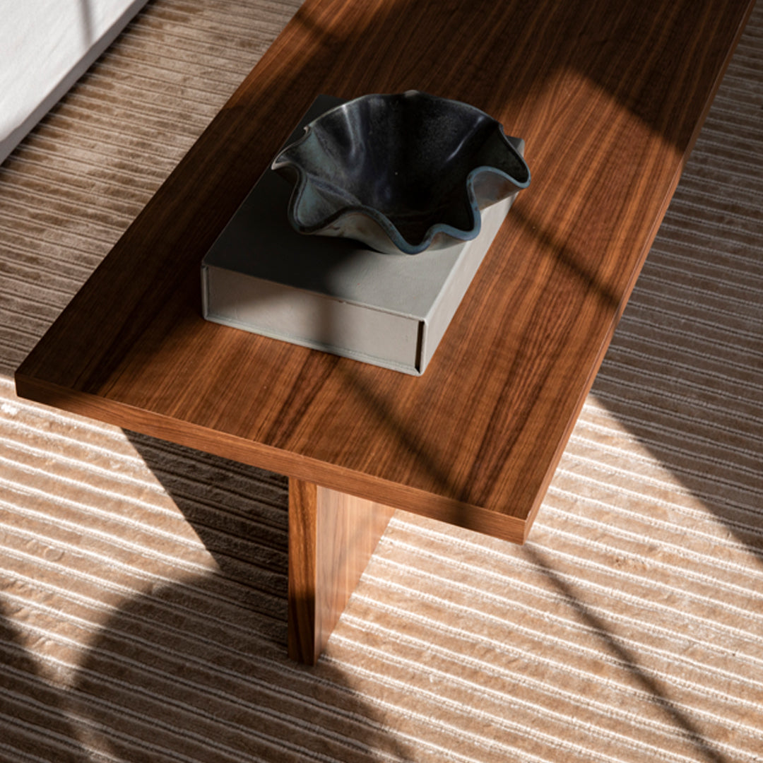 Brown wood coffee table with a smooth finish, gray box, and black decorative bowl on striped beige rug.