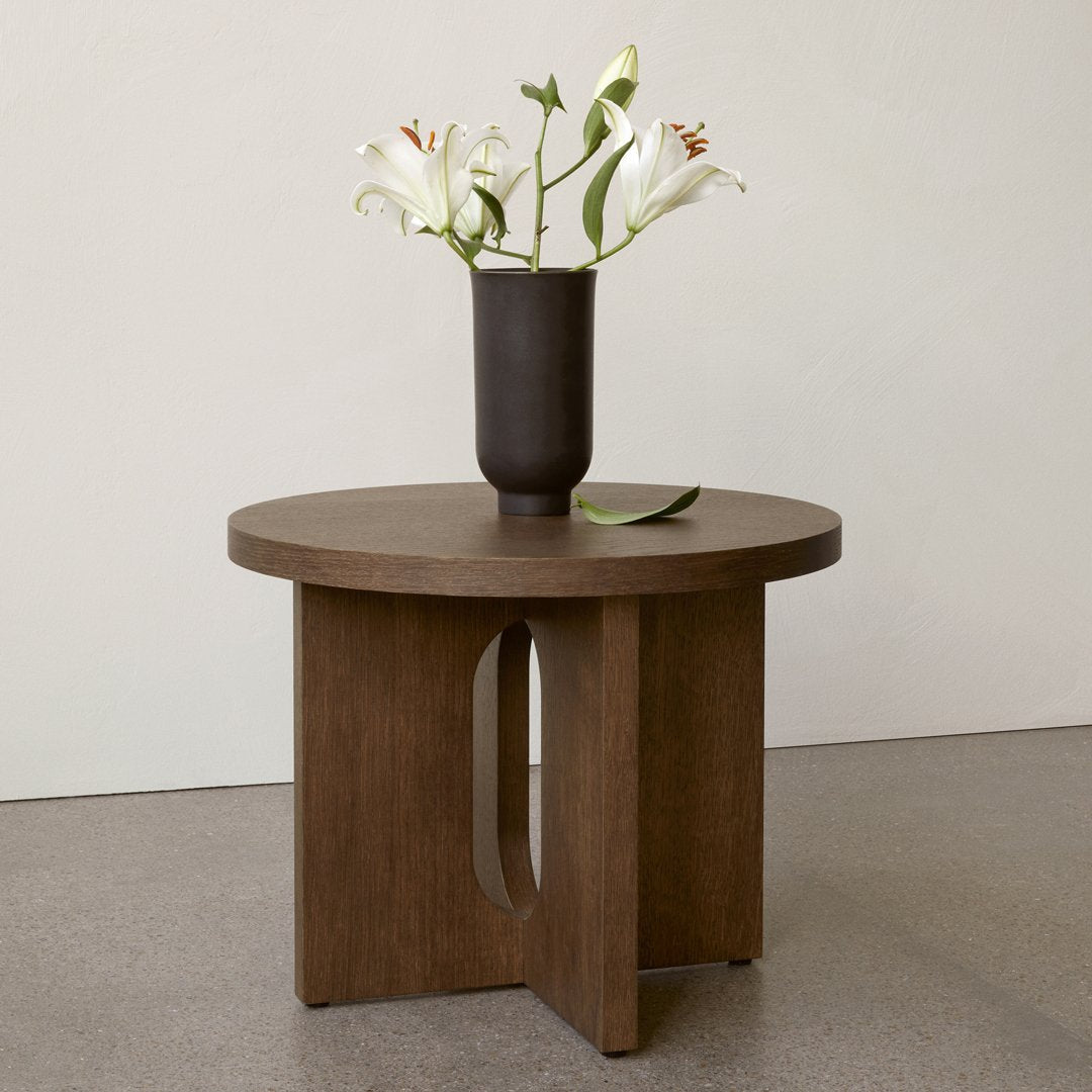 Round wooden side table in rich brown with a minimalist design and subtle cutout pattern, featuring a dark vase and white flowers.