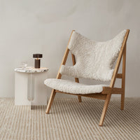 White sheepskin chair on light wooden frame, with a textured cream rug and minimalist side table in a neutral setting.
