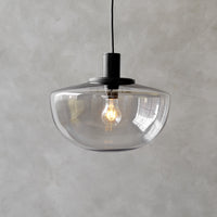 Clear glass pendant lamp with a minimalist black base and warm light bulb, set against a textured gray background.