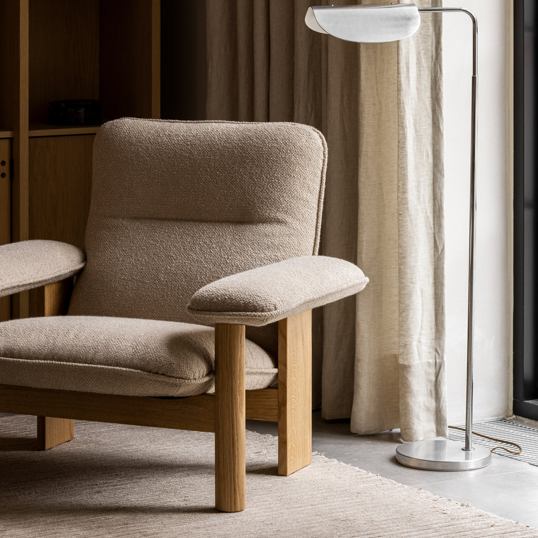 Beige upholstered armchair with light wood legs, positioned beside a silver floor lamp, against neutral-toned curtains.