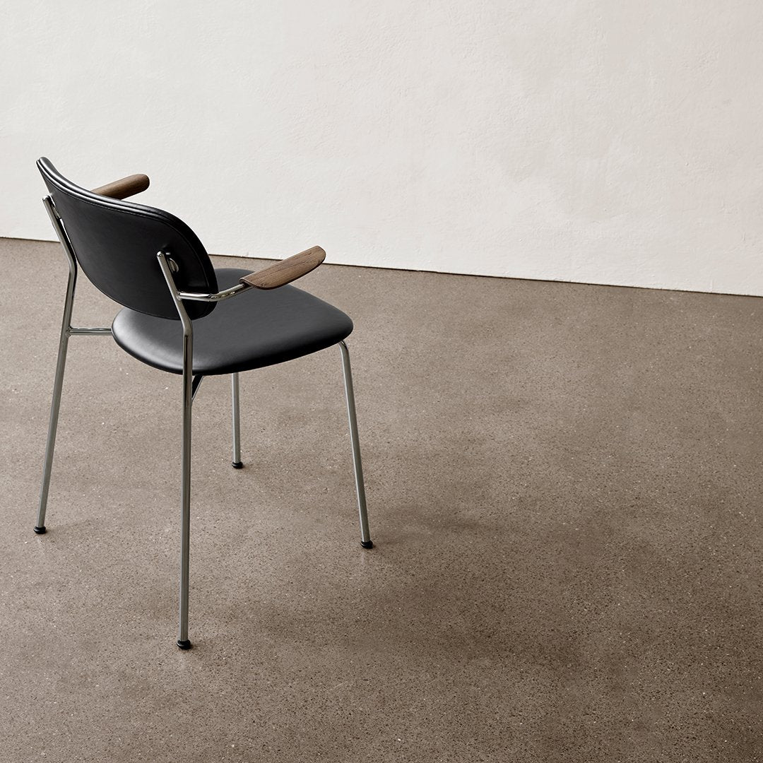 Black chair with wooden armrests, set against a light beige backdrop and dark textured floor.