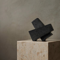 Black geometric sculpture on a beige marble base, featuring a textured surface against a muted gray backdrop.