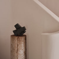 Black geometric sculpture on a rustic wooden pedestal, against a soft beige background.