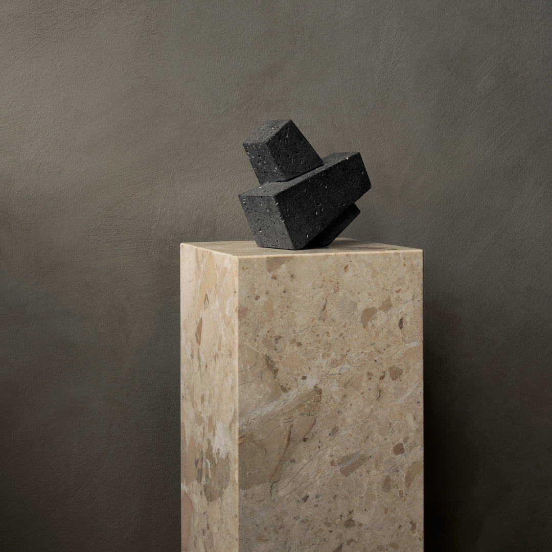 Black geometric shapes atop a textured light stone pedestal, against a muted gray backdrop.