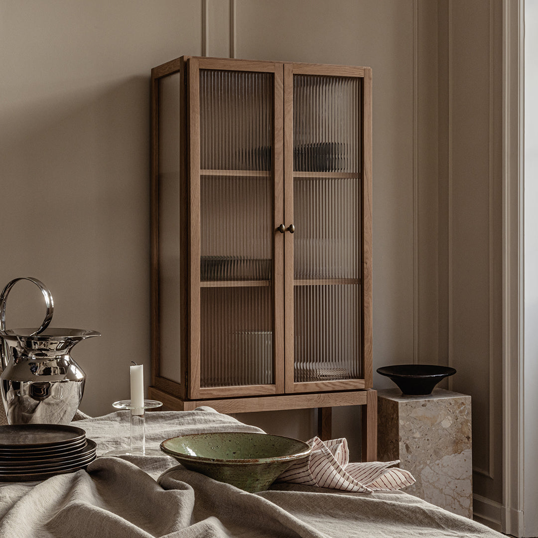 "Light wood cabinet with glass doors, featuring earthy tones and minimalist design, complemented by a neutral backdrop."