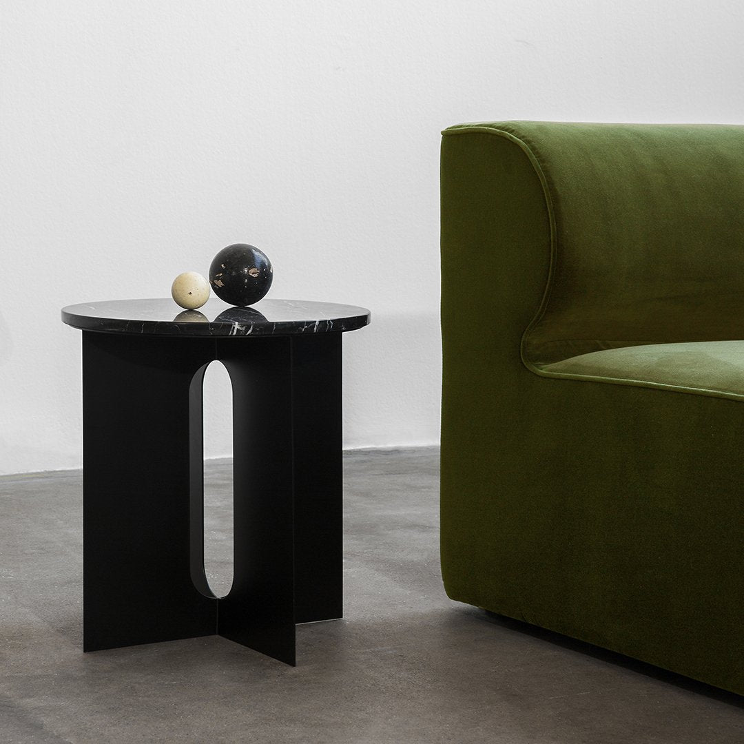 Black round side table with geometric cutout next to a lush green sofa, accented by decorative orbs.