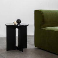 Black round side table with geometric cutout next to a lush green sofa, accented by decorative orbs.