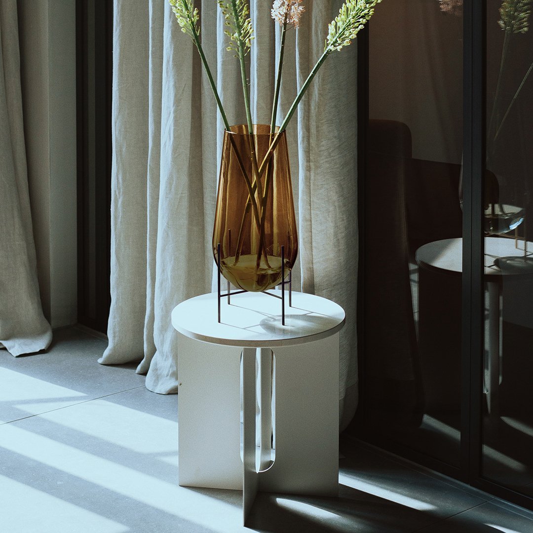 "Modern white side table with an amber vase and green stems, featuring soft shadows and natural light."