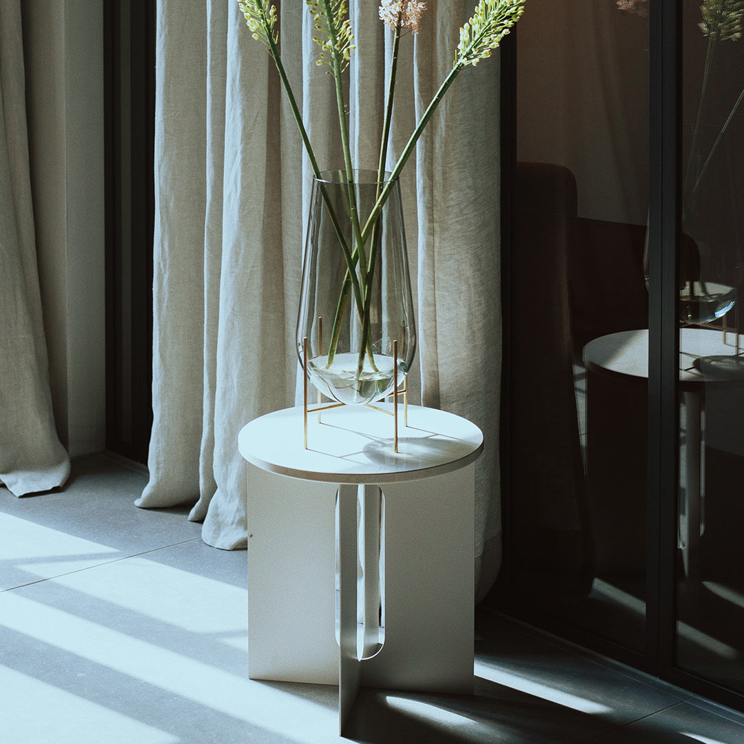 White round side table with a minimalist design, featuring sleek lines and soft shadows against neutral curtains.