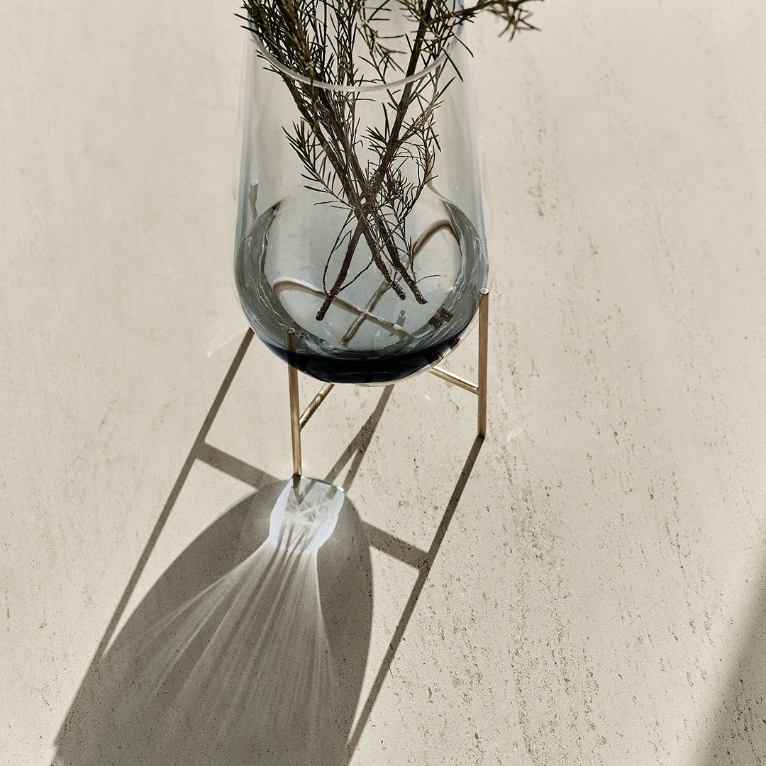 "Clear glass vase with black base, minimalist gold stand, shadows on a light textured surface."