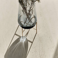 "Clear glass vase with black base, minimalist gold stand, shadows on a light textured surface."