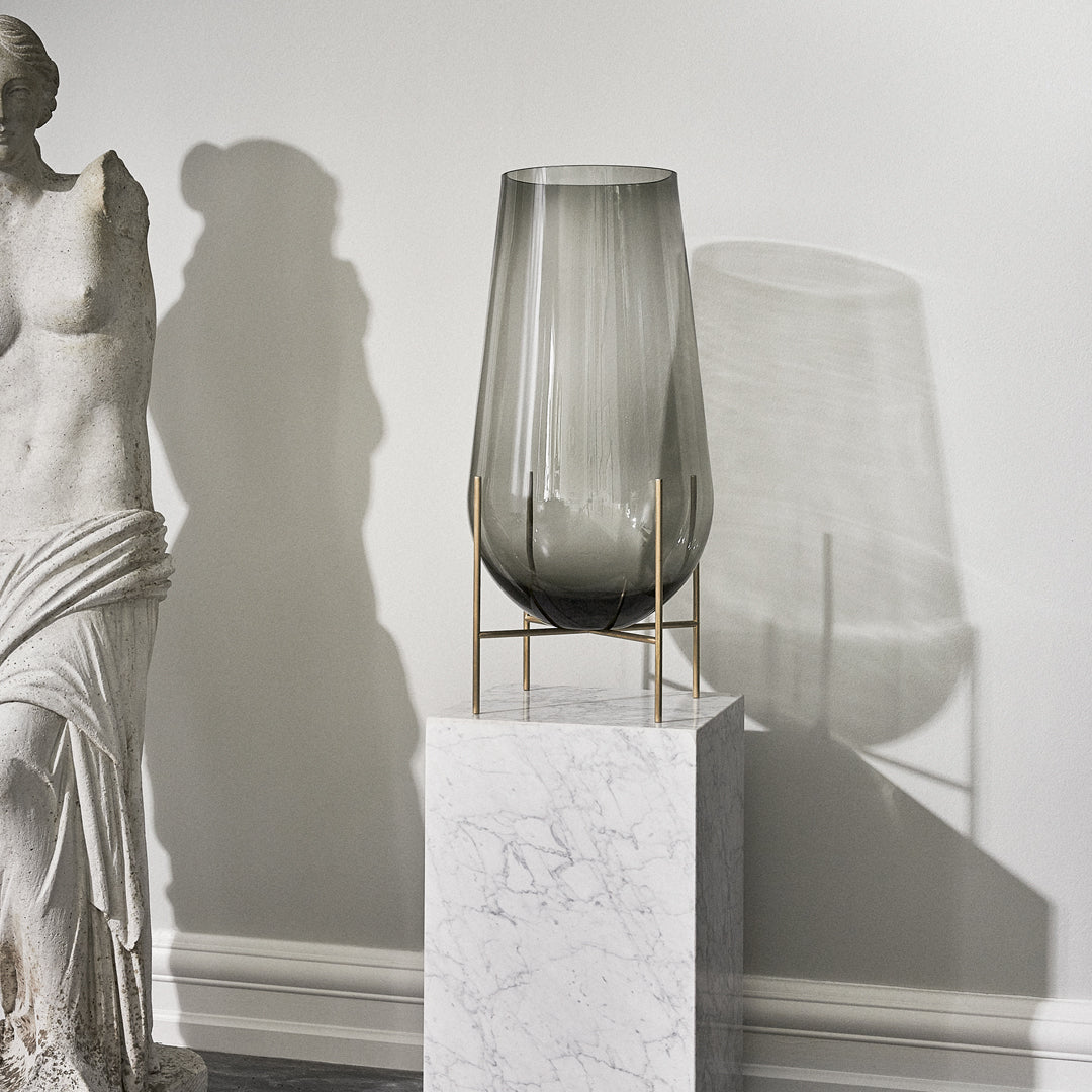 Smoky grey glass vase with vertical stripes on a marble pedestal, creating elegant shadows.