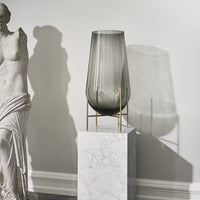 Smoky grey glass vase with vertical stripes on a marble pedestal, creating elegant shadows.