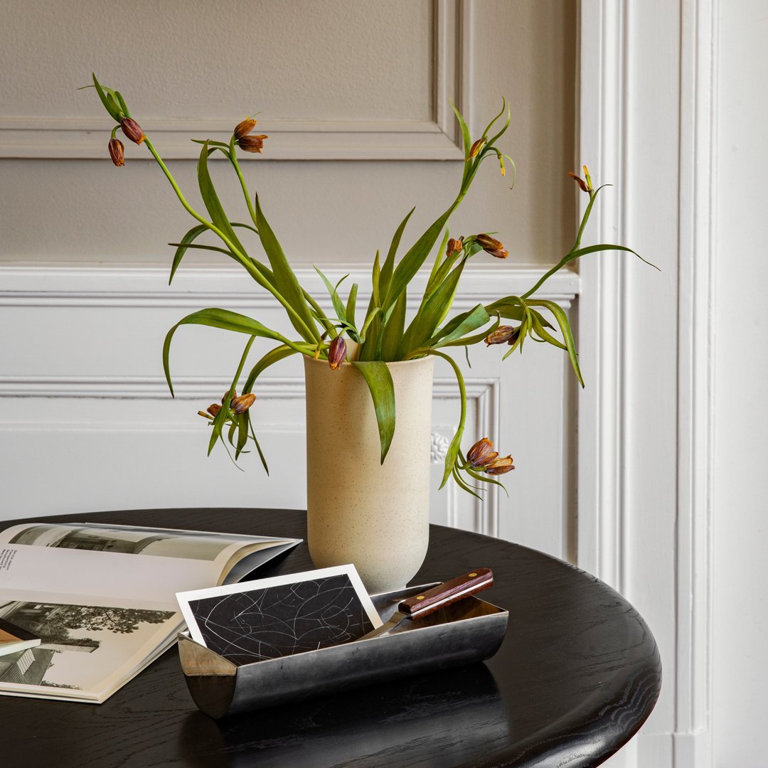 Vase with earthy tones, adorned with green leaves and brown flower buds, set against a neutral background.