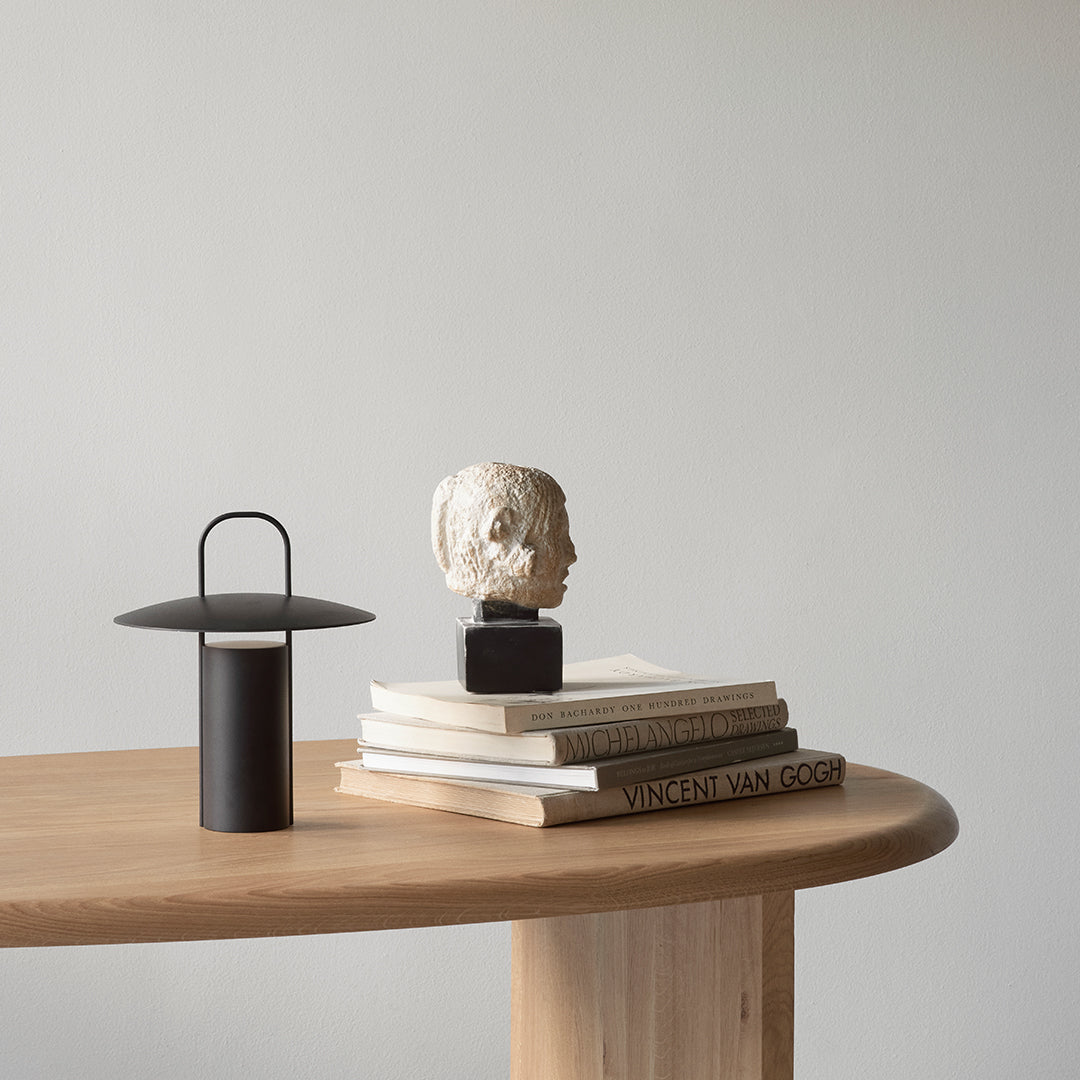 Black lamp atop books with cream, beige, and brown tones, featuring a white sculptural head accent against a light gray background.