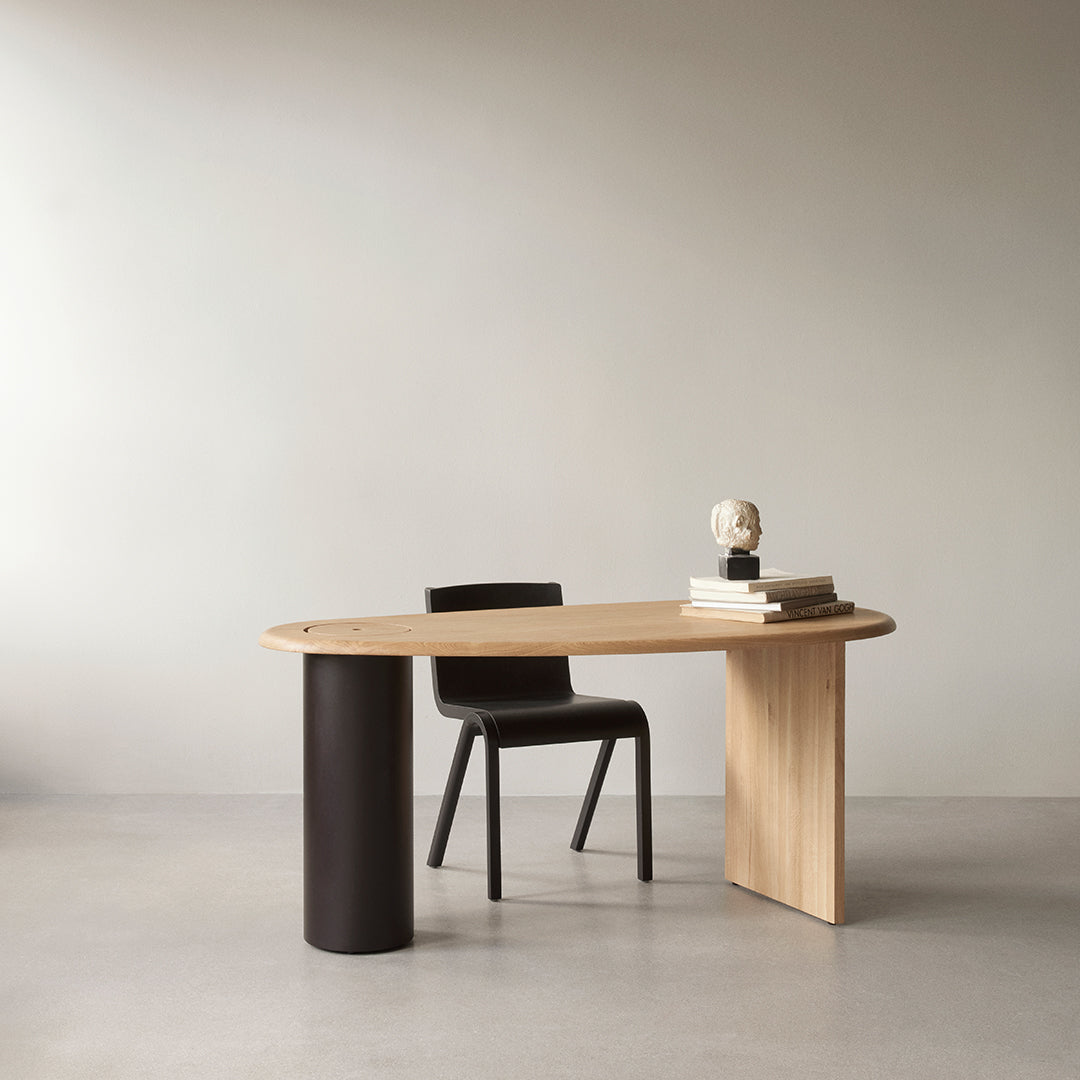 Natural wood desk with a light oak finish, paired with a matte black chair on a neutral-toned floor. Modern design.