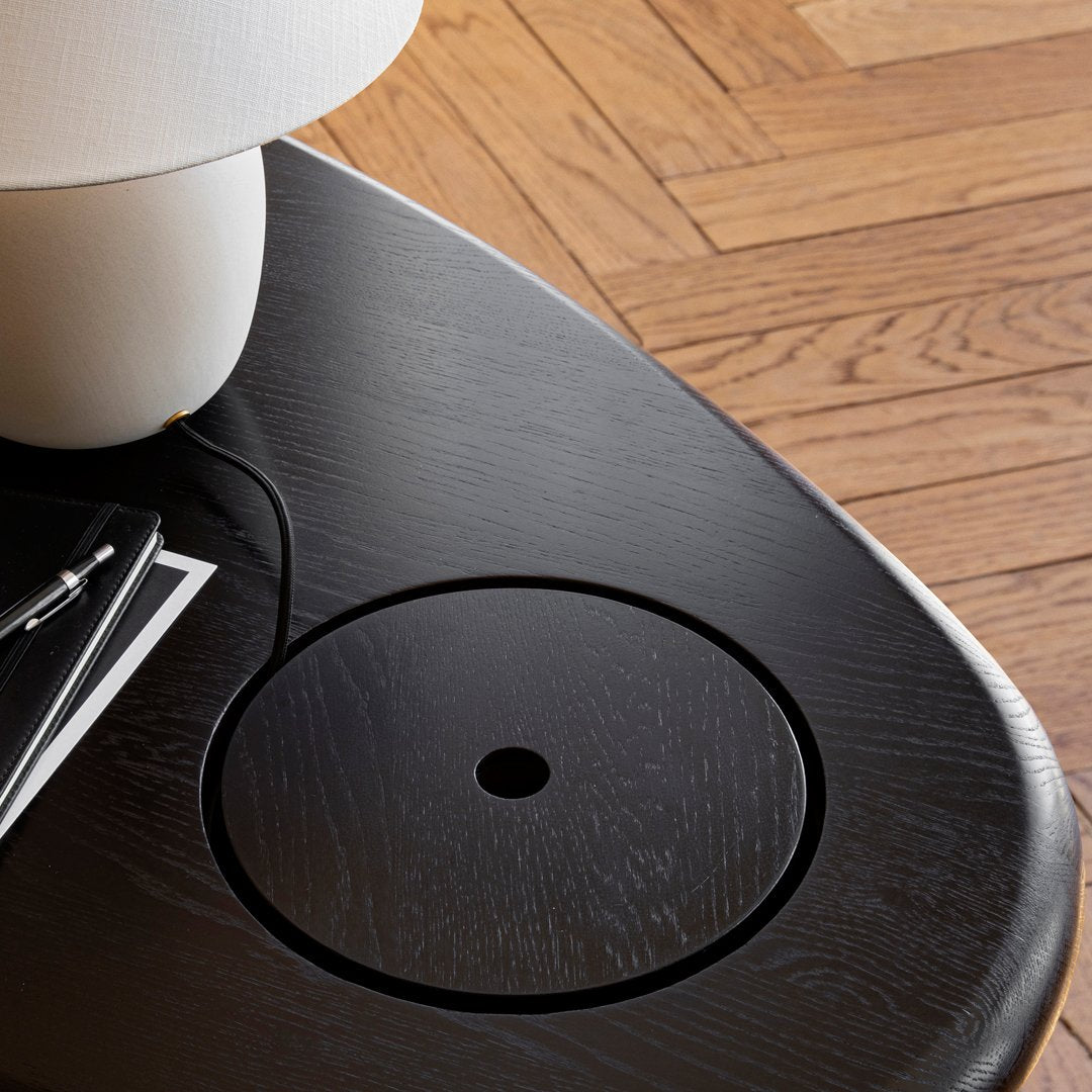 Black circular tabletop with subtle wood grain pattern, complemented by a sleek white lamp and dark wood accents.