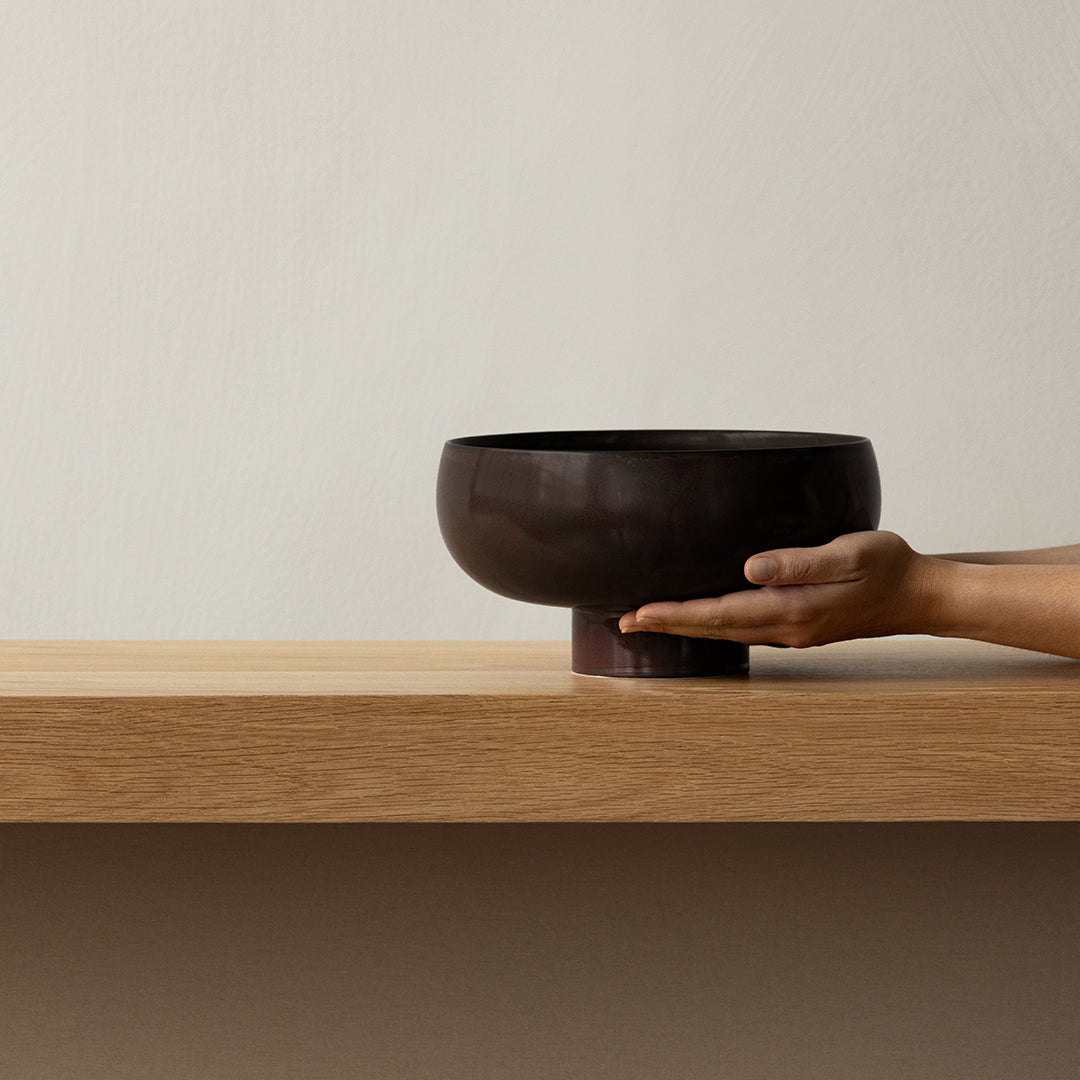 Dark brown ceramic bowl with a smooth, cylindrical shape, displayed on a wooden shelf.