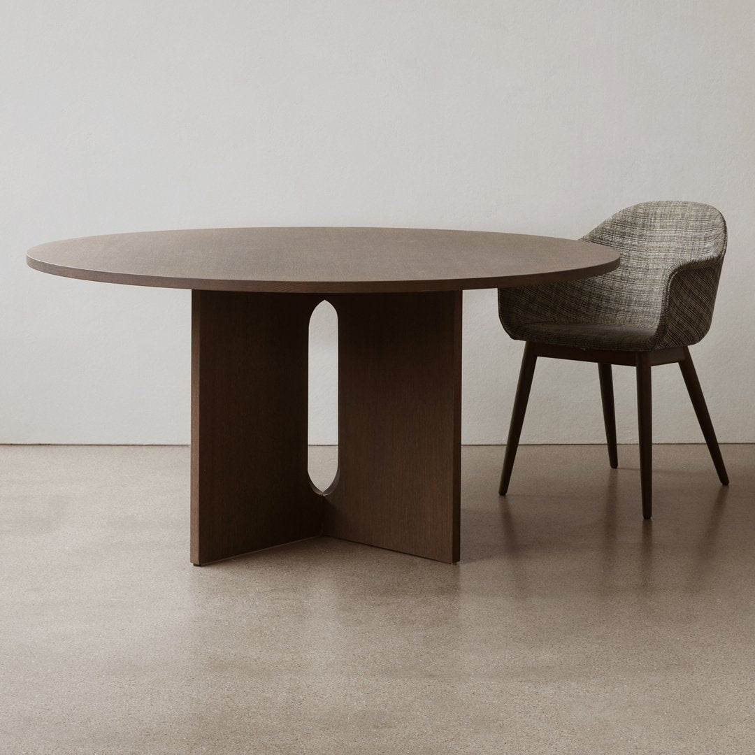 Round wooden dining table with warm brown tones and a unique cutout base, complemented by a textured gray chair.